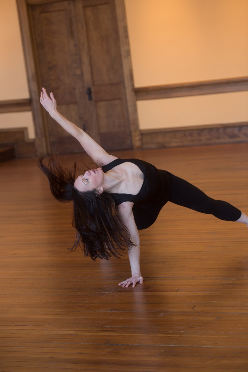 A dancer is in a lunge with one arm supporting her on the ground and one hand reaches for the sky. Her long brown hair flies in response.